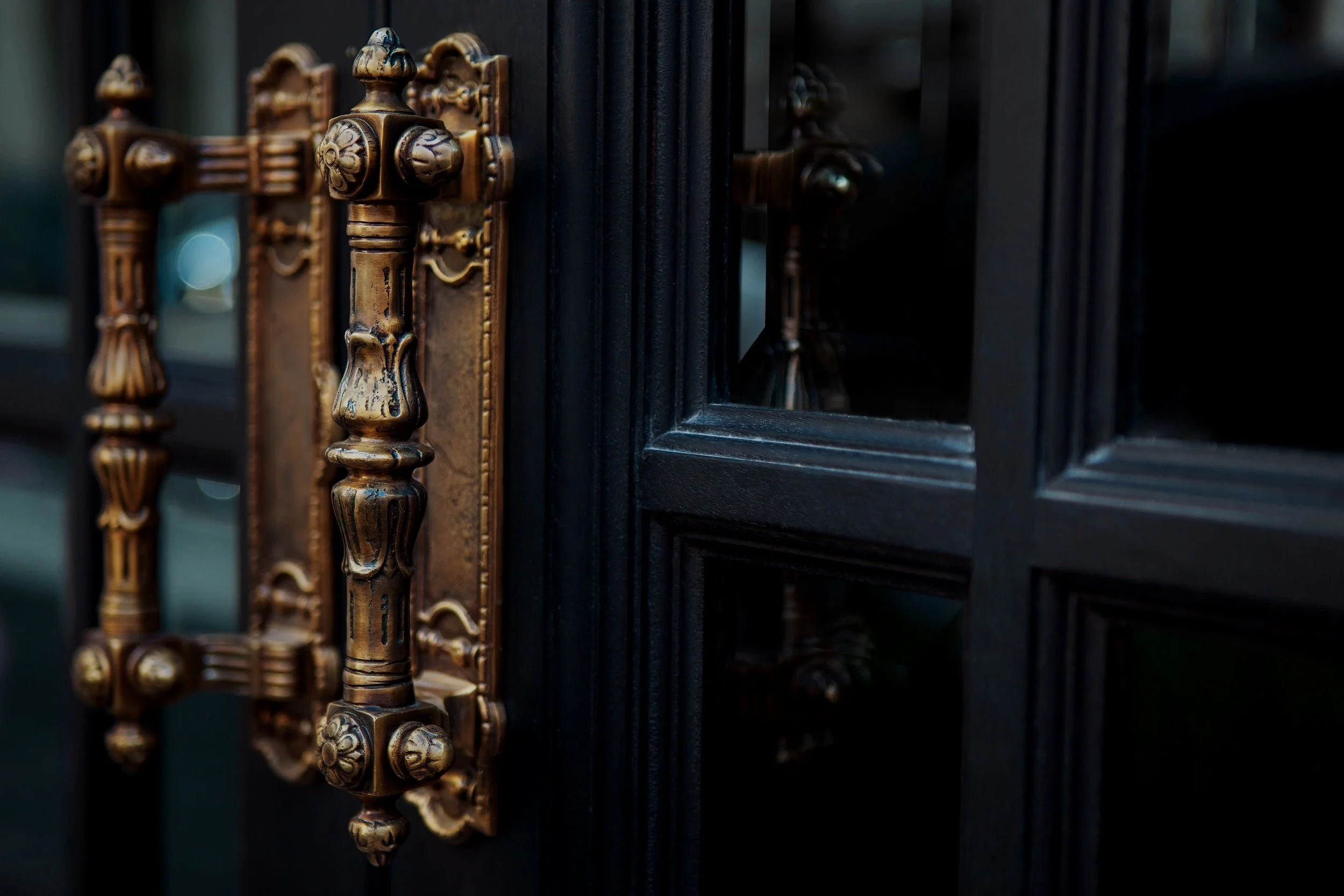 Close-up of an ornate, vintage brass door handle and lock on a dark wooden door with glass panels.