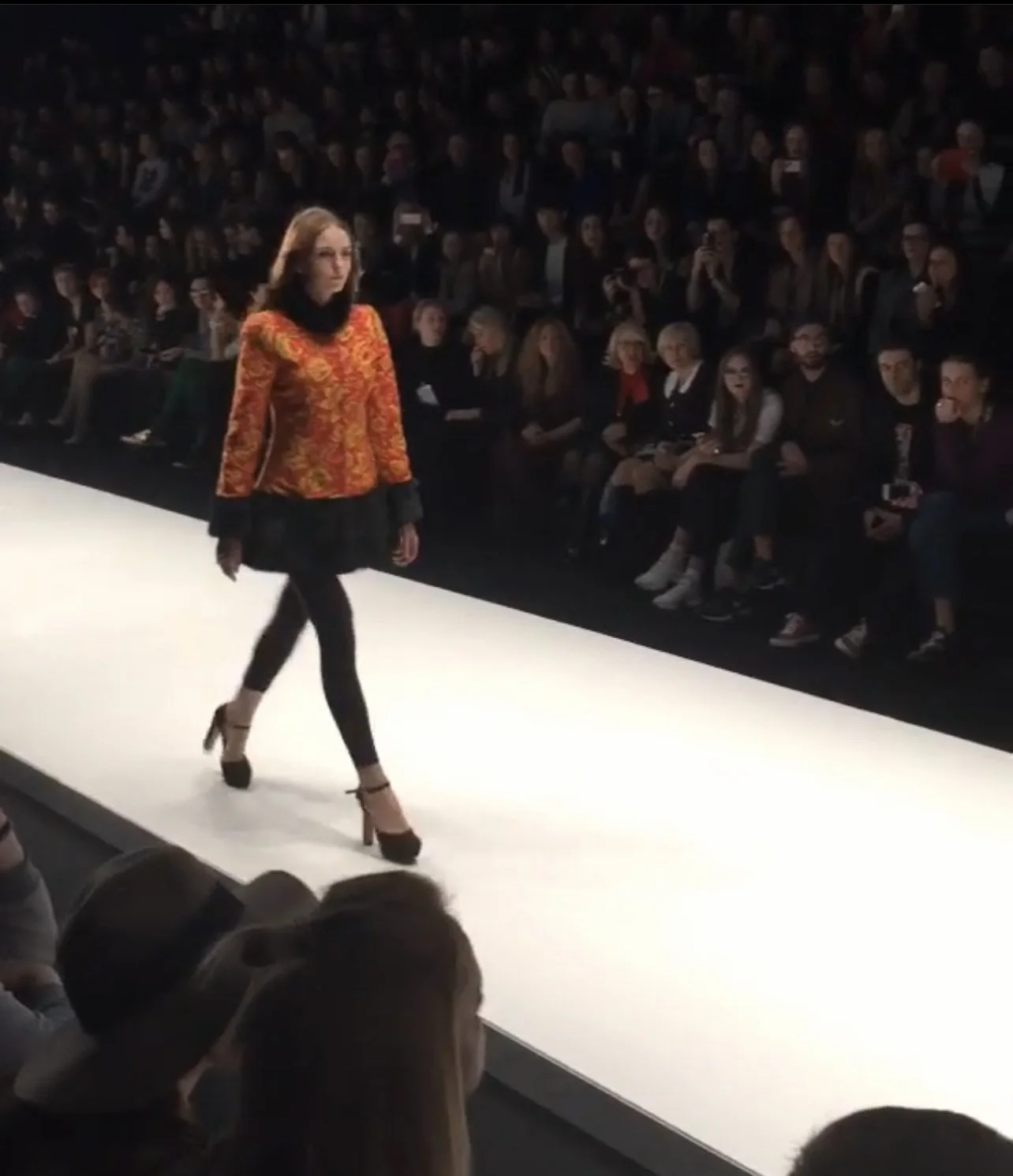 A female model walking on a runway at a fashion show, wearing a colorful patterned jacket, a black skirt, black tights, and high heels, with an audience watching in the background.