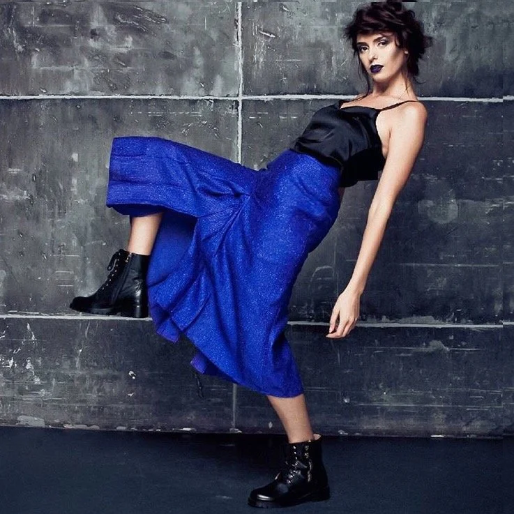 A woman with dark hair and makeup wearing a black top, a blue skirt, and black combat boots, leaning against a concrete wall.