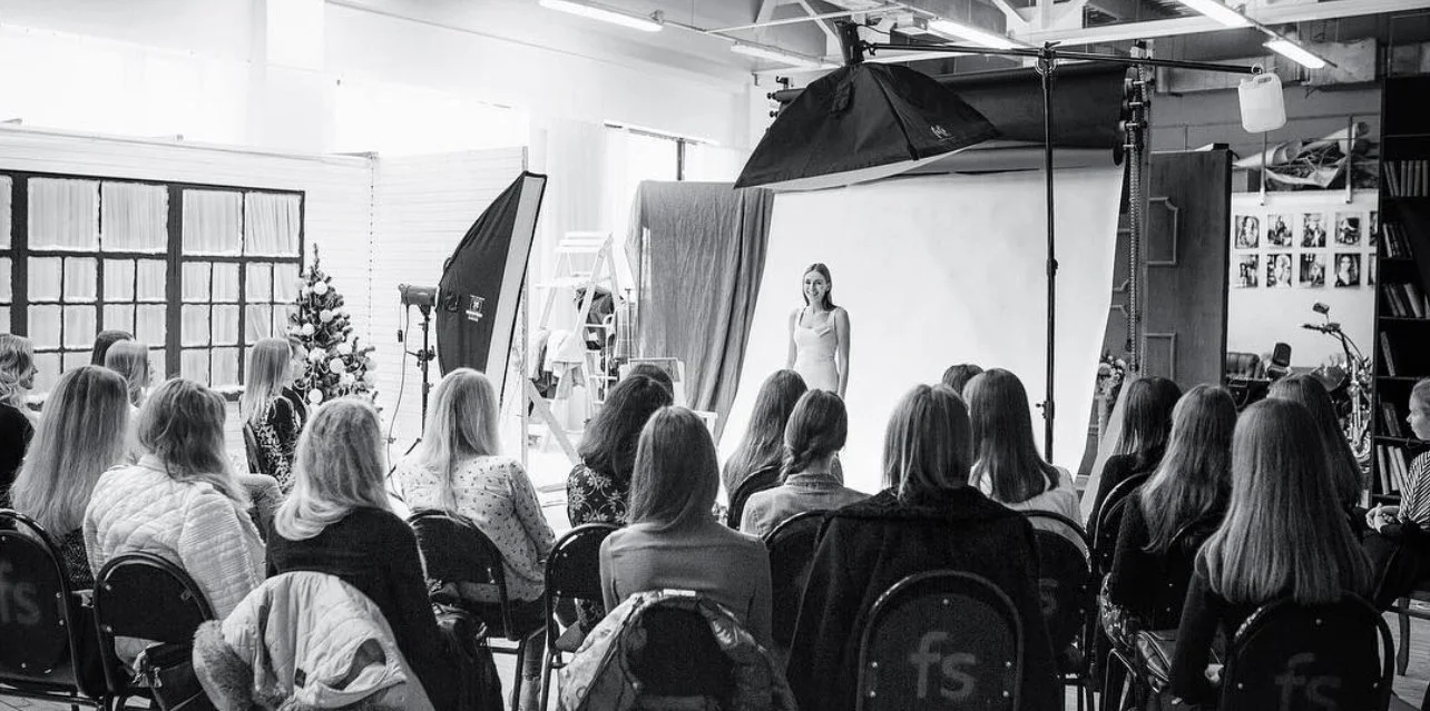 A black-and-white photo shows a group of women seated in a photography studio, watching a woman model in front of a white backdrop. Studio lights and equipment are visible, and a Christmas tree is in the background.