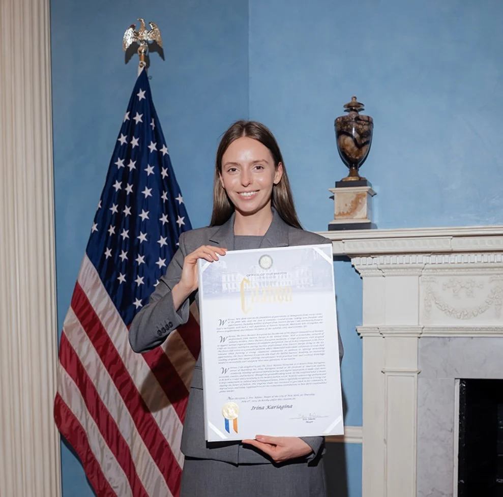 A woman in a gray suit holding a large certificate, standing in front of an American flag and a blue wall with a fireplace.