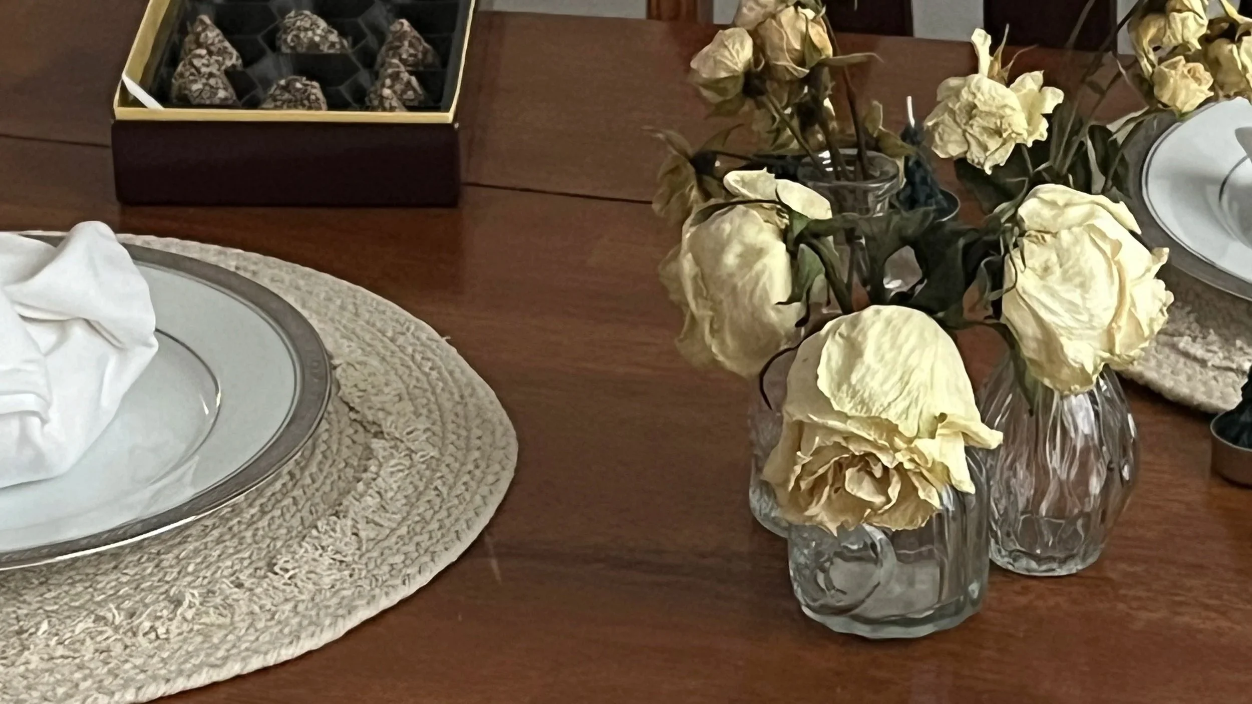 Dried white roses in two textured glass vases on a wooden dining table, with a placemat, white napkin, and a tray of chocolates in the background.