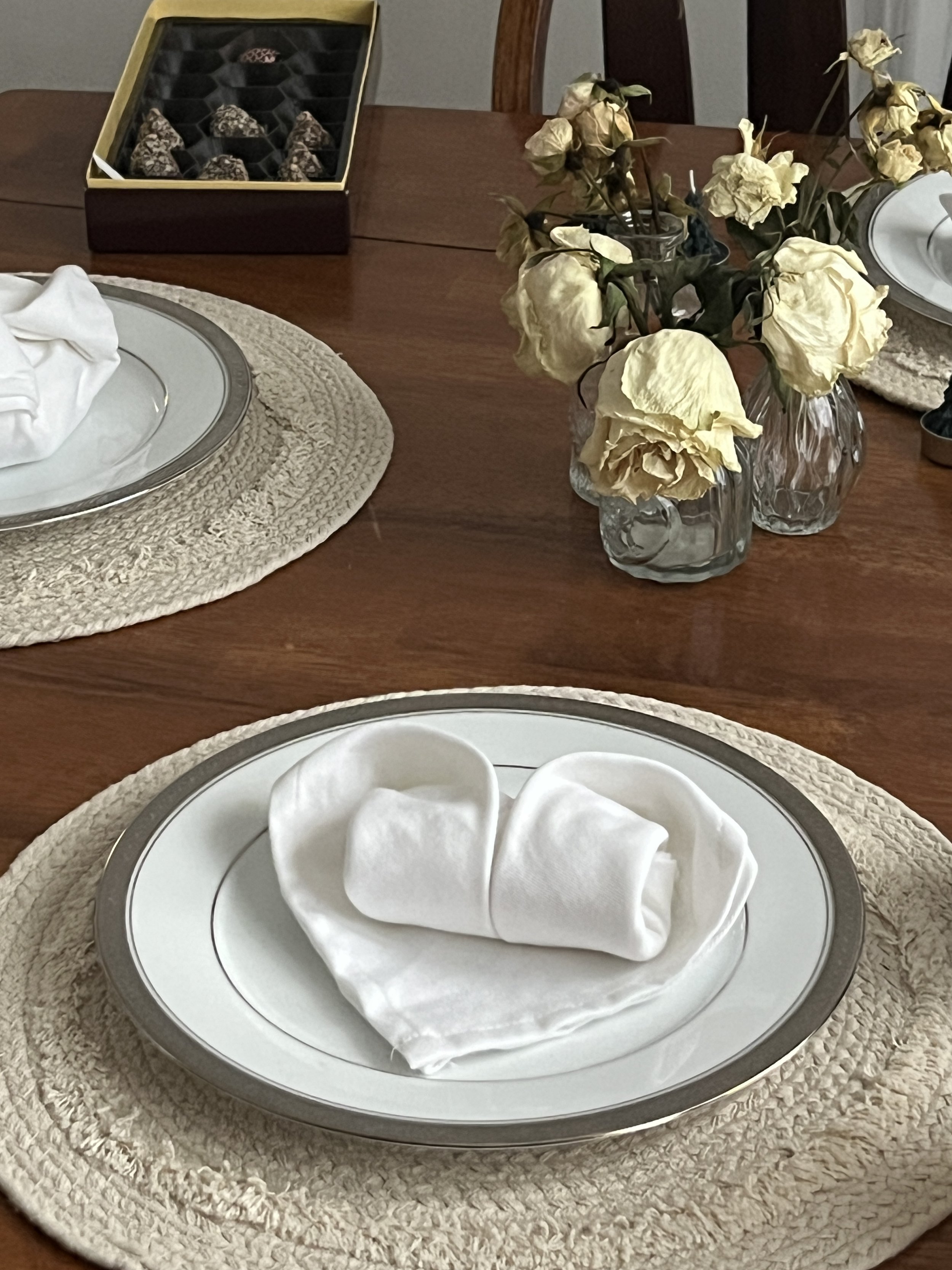 A table setting with a white plate, a neatly folded white napkin, and a beige woven placemat. In the background, there are two glass vases with wilted yellow roses and a box of assorted chocolates on a dark wooden table.