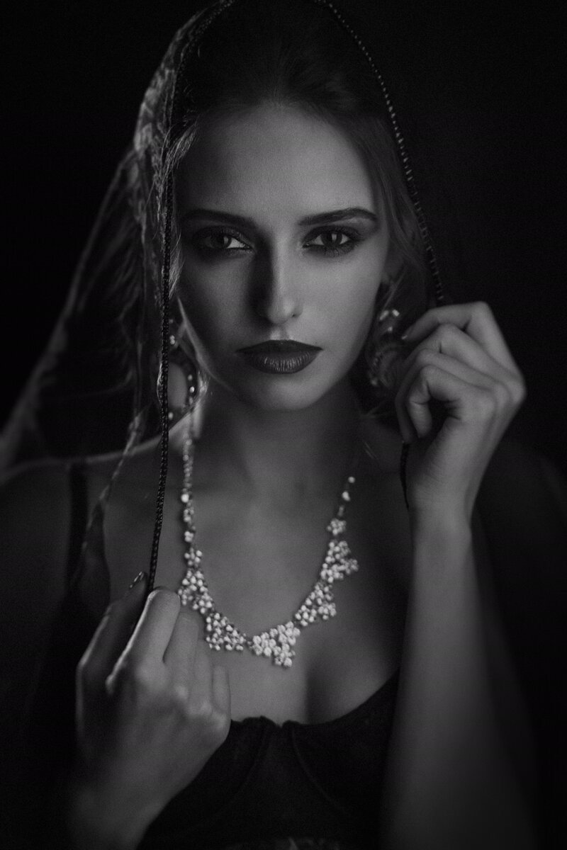 Black and white portrait of a woman wearing a headscarf, earrings, and a necklace, with a serious expression, looking directly at the camera.