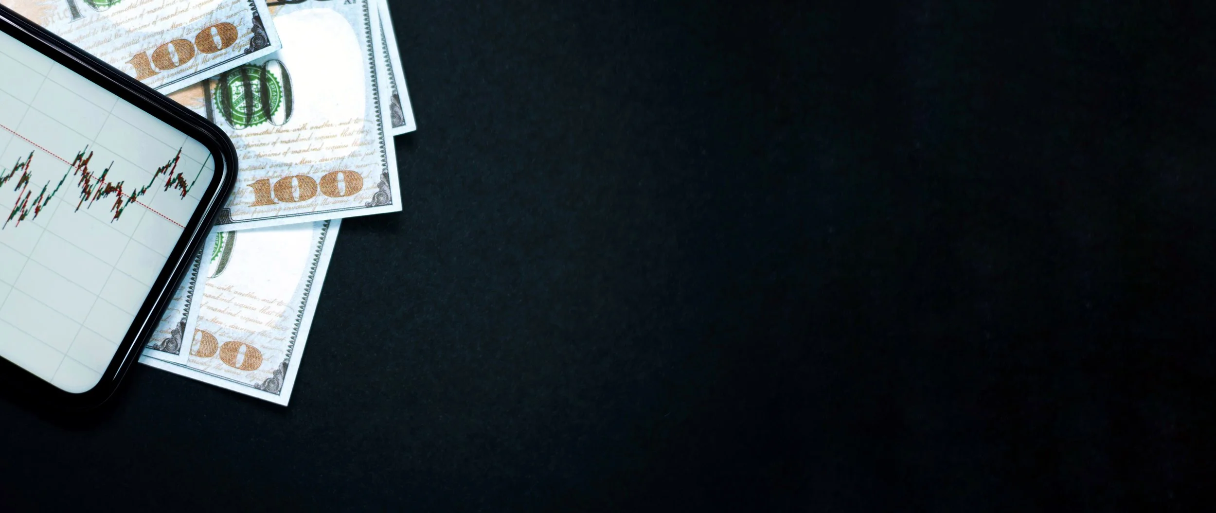 Smartphone displaying a stock market graph placed on a dark surface alongside a few US hundred-dollar bills.