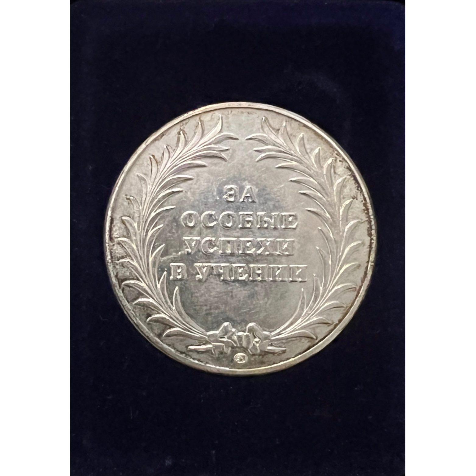 A silver coin with engraved laurel branches and a ribbon at the bottom, featuring a Cyrillic inscription in the center.
