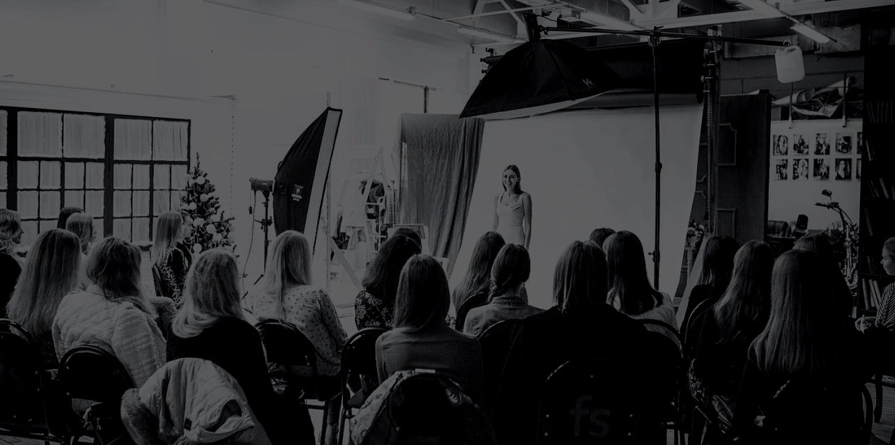 A photography studio with a woman modeling on a white backdrop, large studio lights, and an audience watching the session.