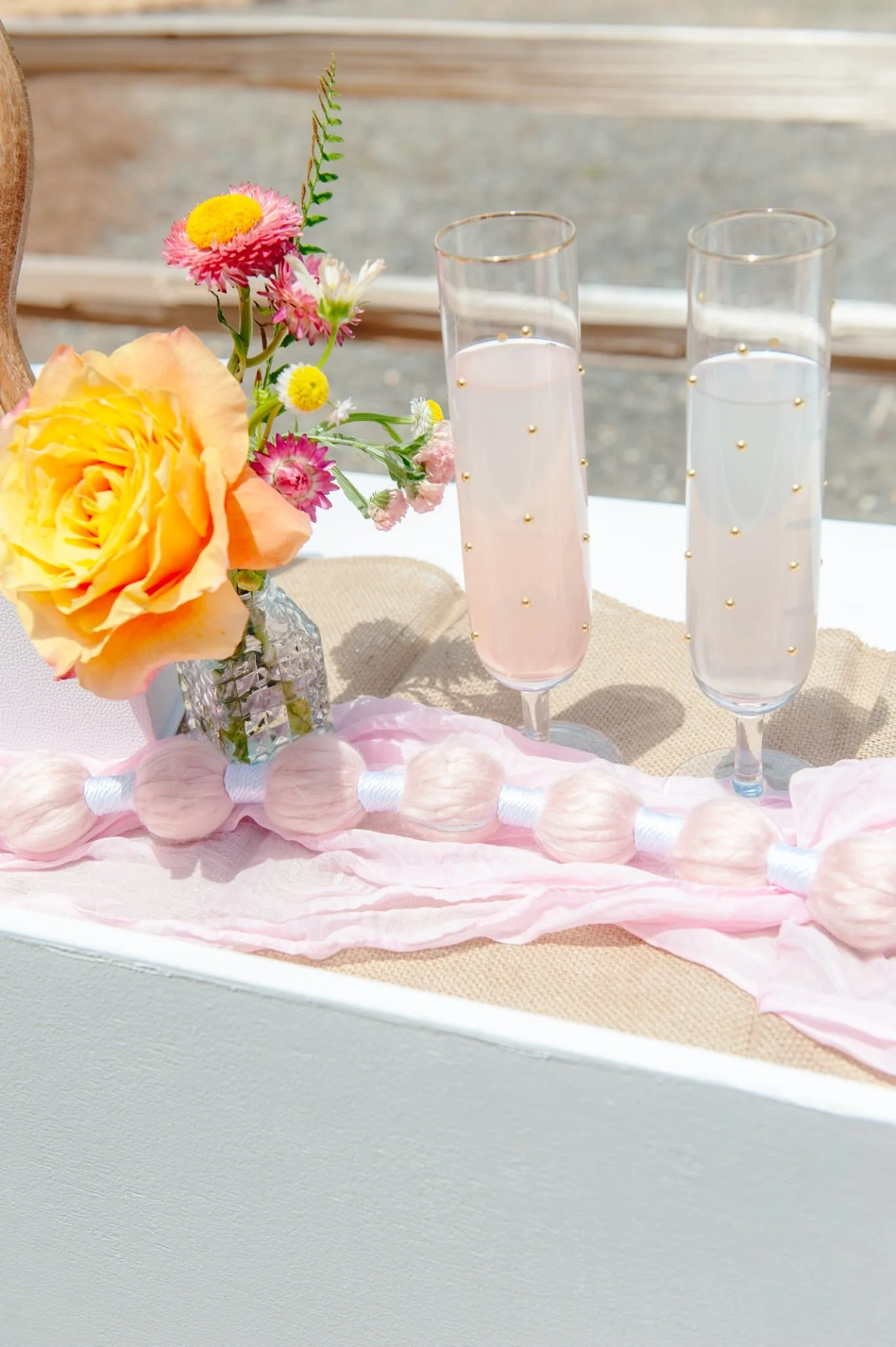 A floral centerpiece with yellow and pink flowers in a crystal vase, two tall champagne flutes with pink and clear liquids, and pink felt ball garland on a table with a linen runner.