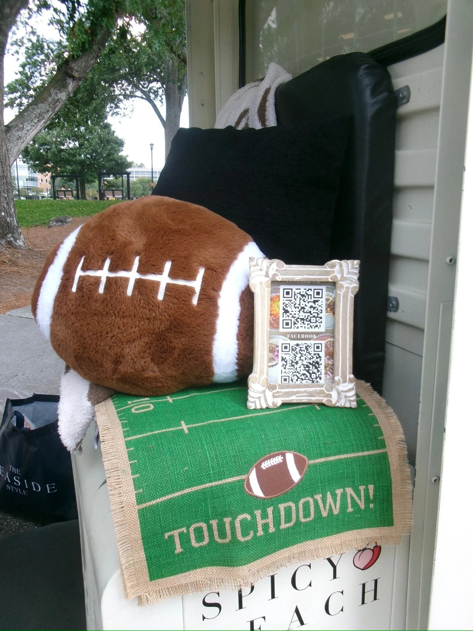 Decorative display with a plush football pillow, a QR code in a frame, a black cushion, a towel, and a blanket with a football theme, set outdoors with trees and benches in the background.