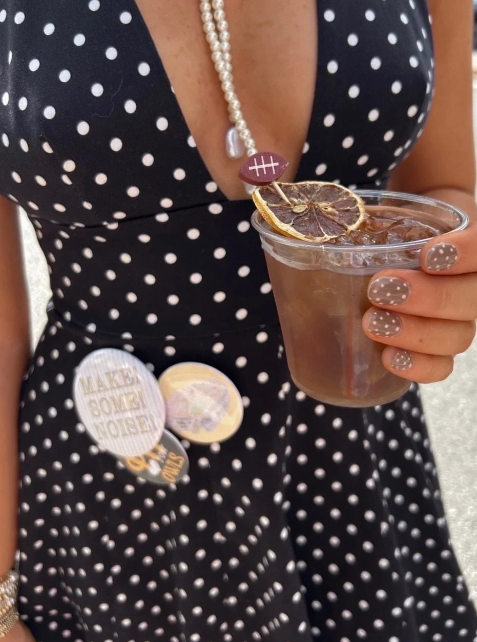 A woman wearing a black dress with white polka dots, a pearl necklace, and matching polka dot nails, holding a plastic cup of iced coffee garnished with a dried lemon slice and a small decorative football pick.
