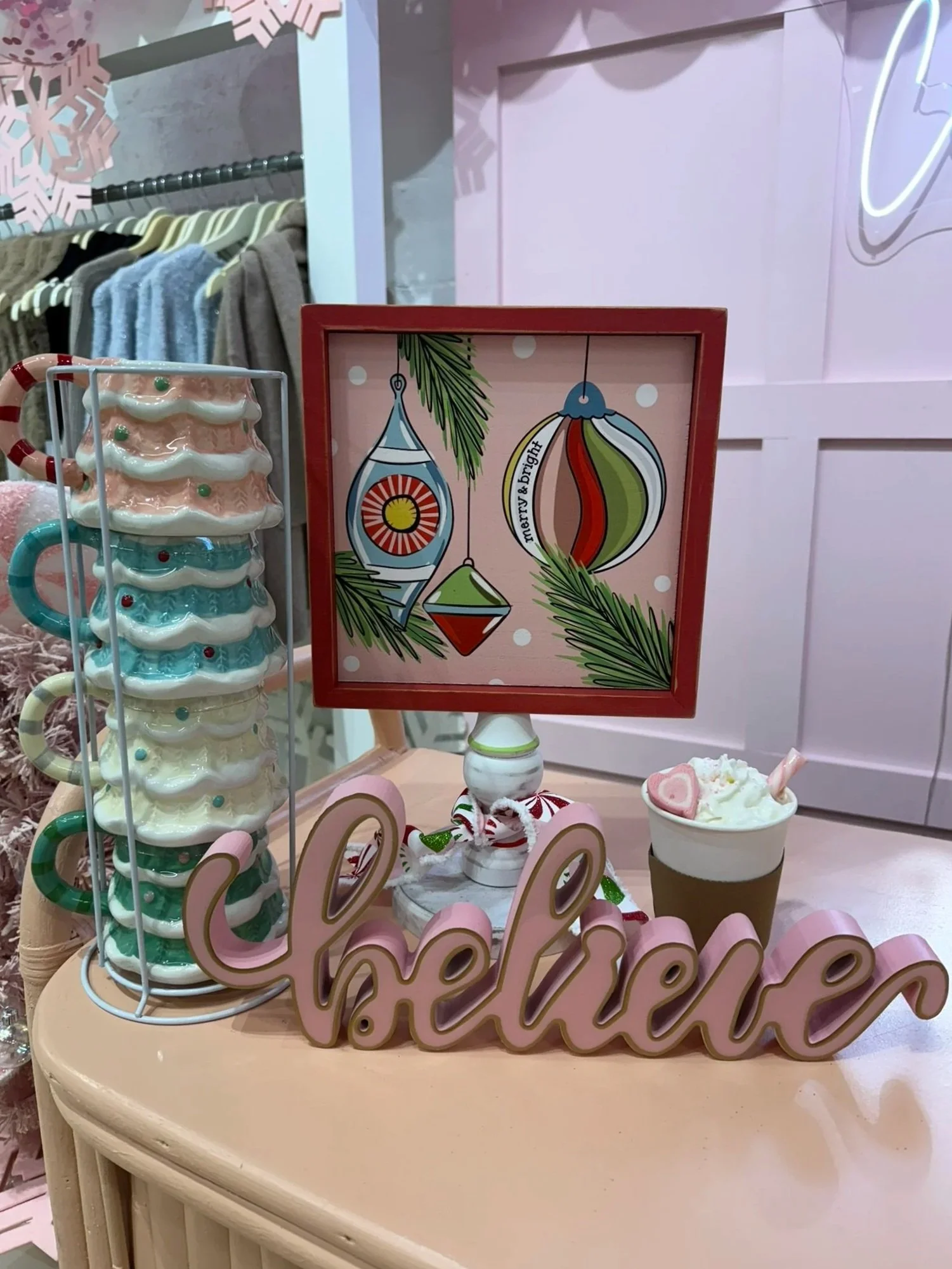 Display with Christmas-themed decorations, including a pink "believe" sign, a mug with whipped cream and a heart-shaped marshmallow, a framed illustration of colorful holiday ornaments, and a ceramic ornament stand with holiday mugs, set on a pink su