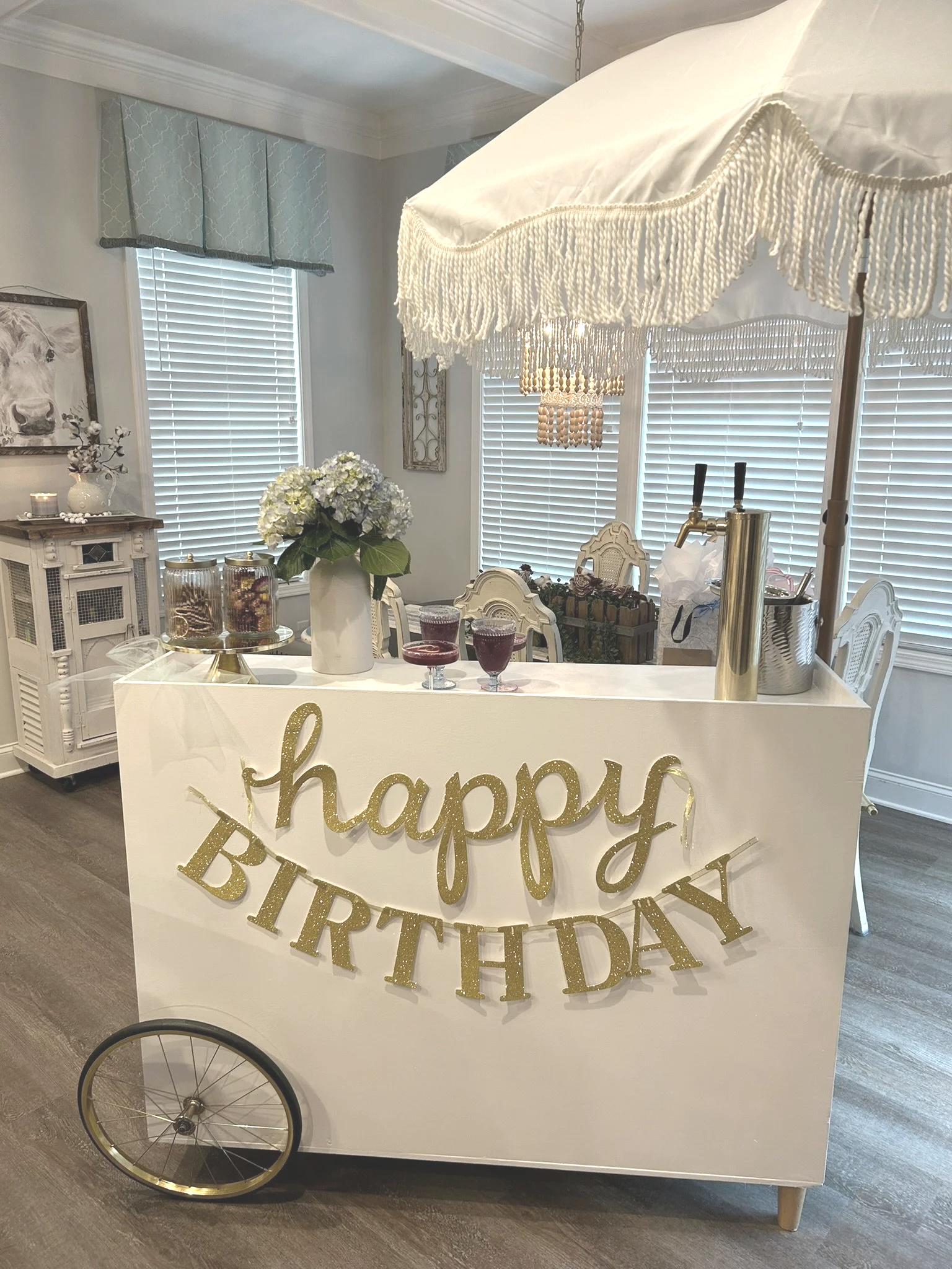 Decorated bar cart with a white canopy, gold and white "happy birthday" banner, flowers, drinks, and party supplies in a warmly lit room.