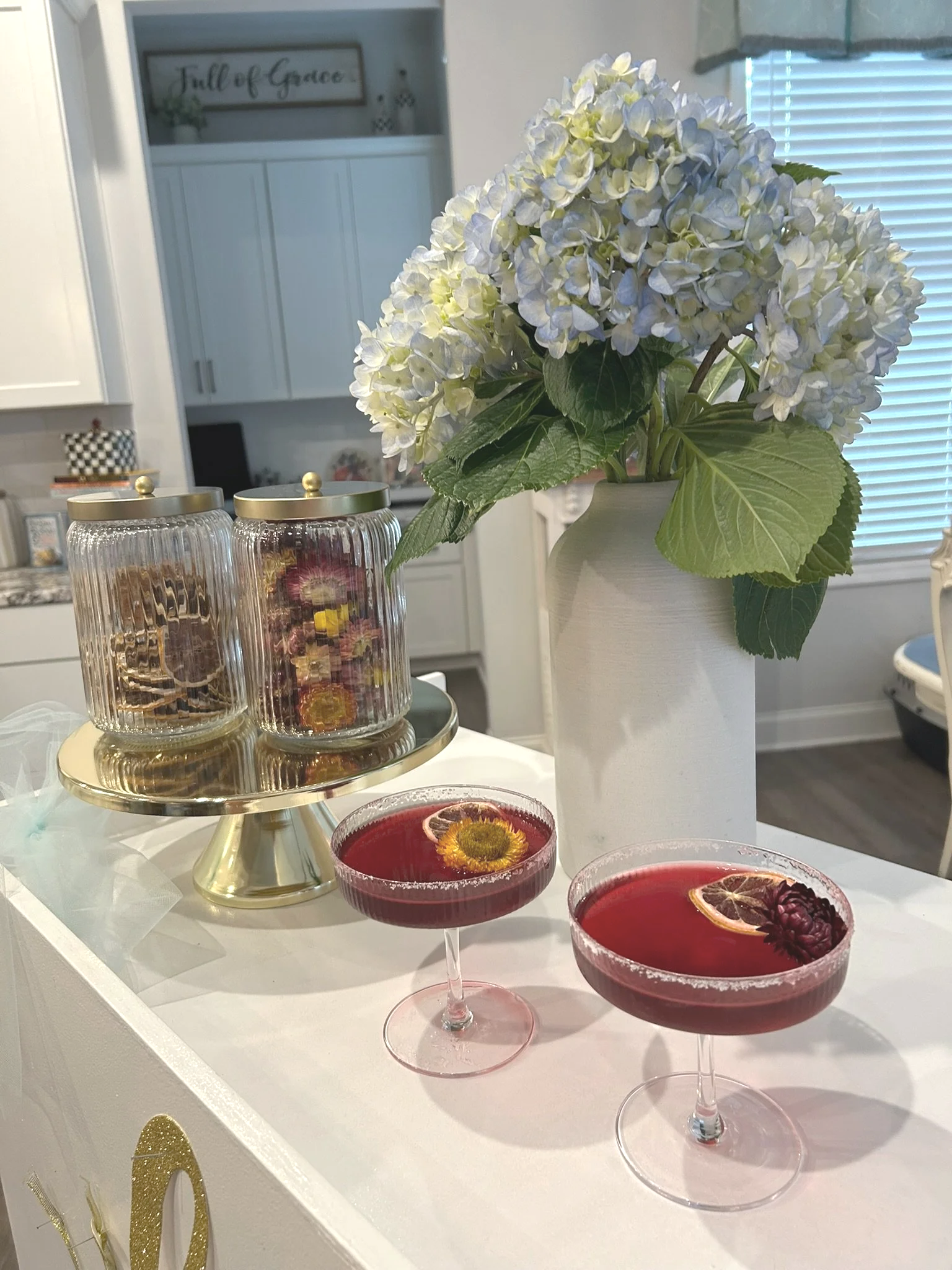 Two pink cocktails with lemon slices and flowers garnish, a white vase with white and green hydrangea flowers, and glass jars with snacks on a gold cake stand, all on a white surface in a kitchen setting.