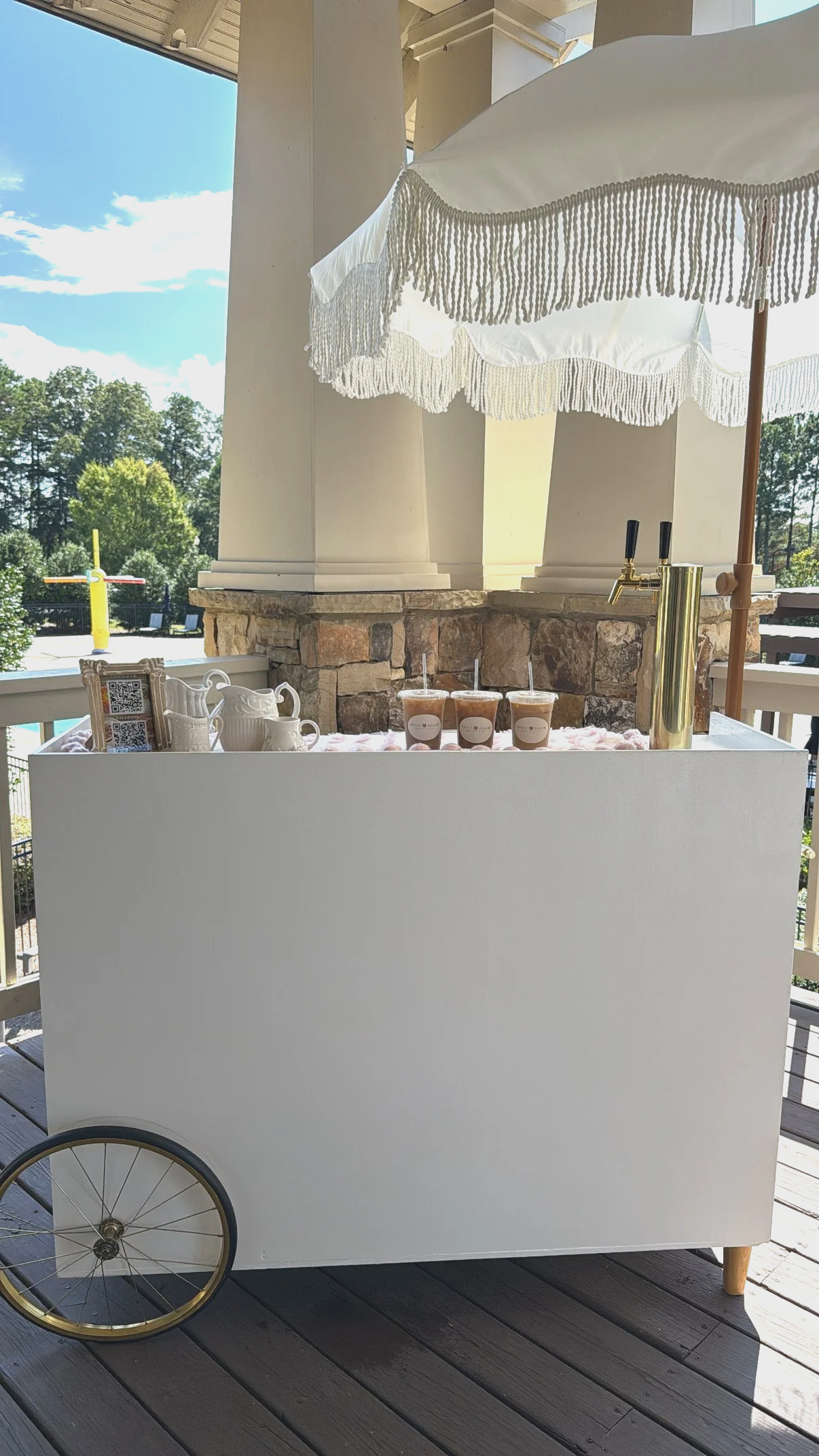 A white mobile beverage cart with a large umbrella, holding three iced drinks, tea set, QR code, and a gold-colored beverage dispenser, on a wooden deck with trees and a pool in the background.
