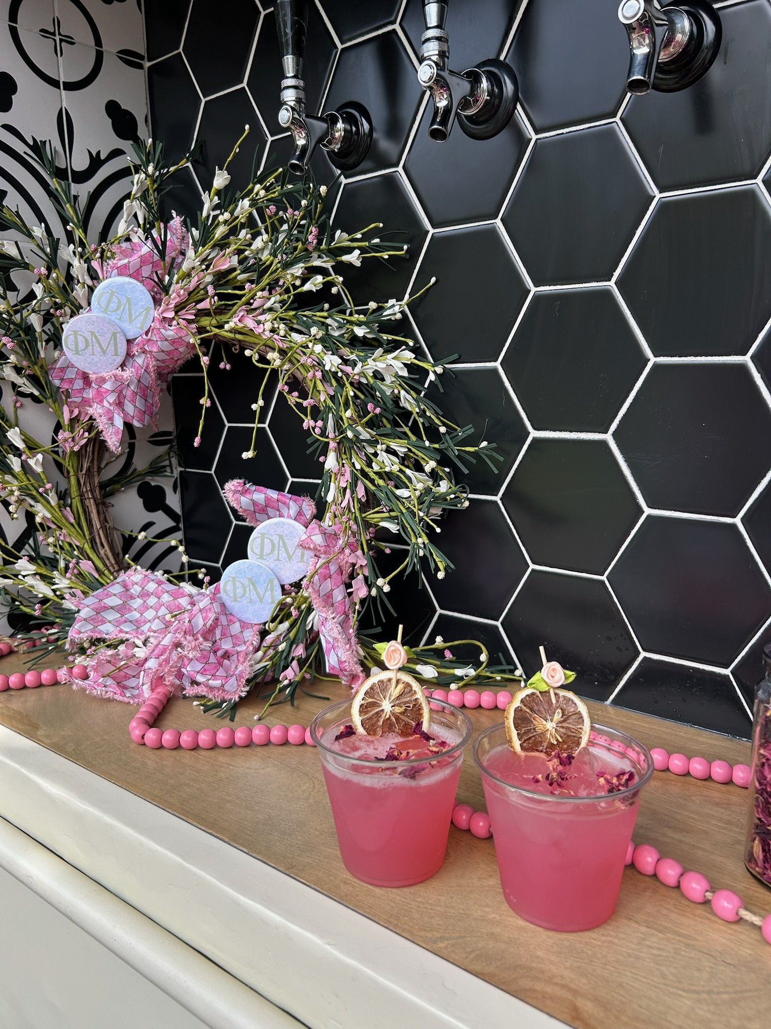 Pink drinks with dried lemon slices and rose petals on a wooden surface, decorated with pink beads and a floral wreath with pink and white flowers against a black hexagonal tile wall.