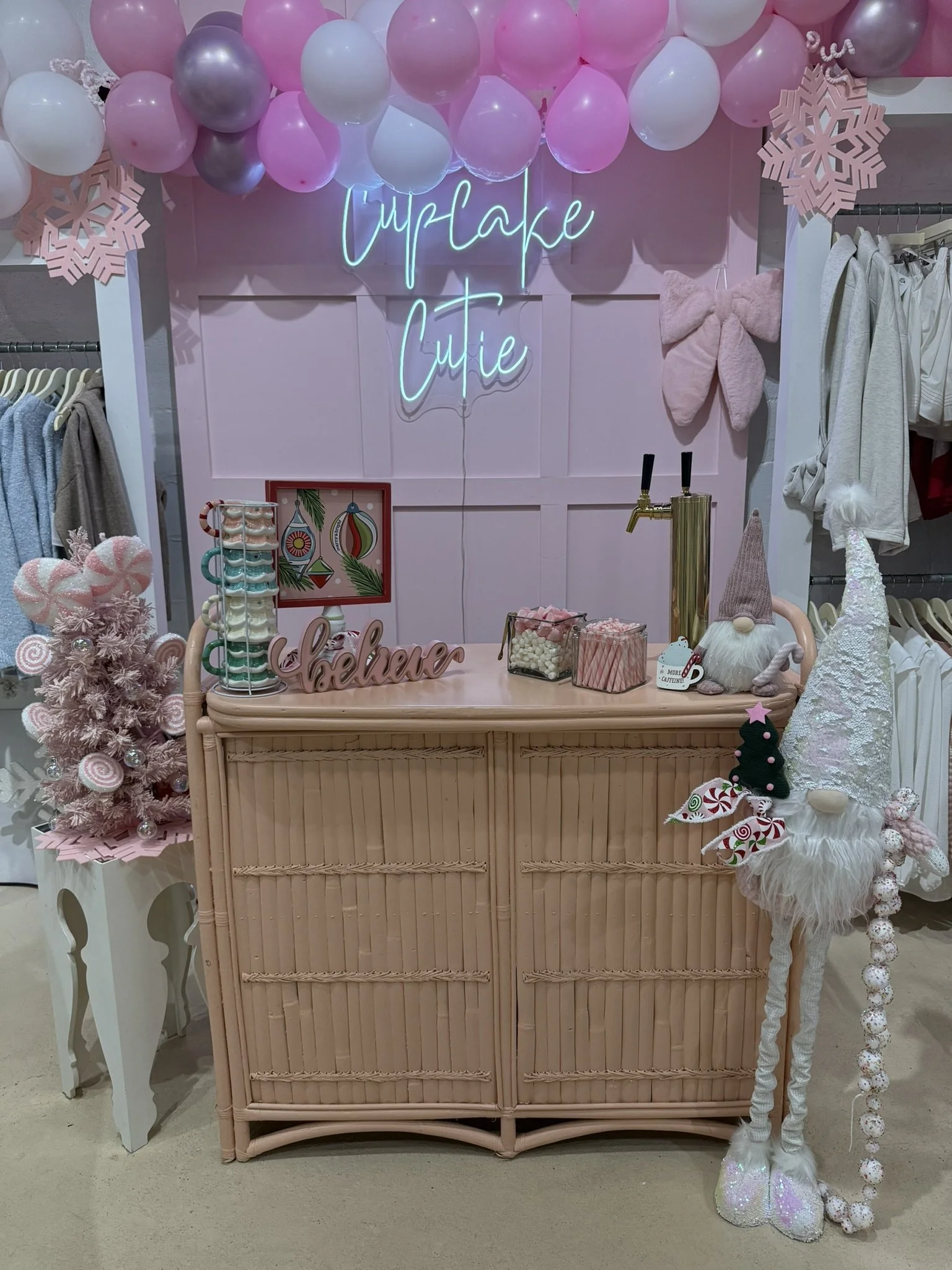 A holiday-themed display with pink, white, and purple balloons, neon sign reading 'Cupcake Cutie,' pink paper snowflakes, and festive decorations including a pink Christmas tree, plush gnome, candy cane decorative items, and a wicker dresser.