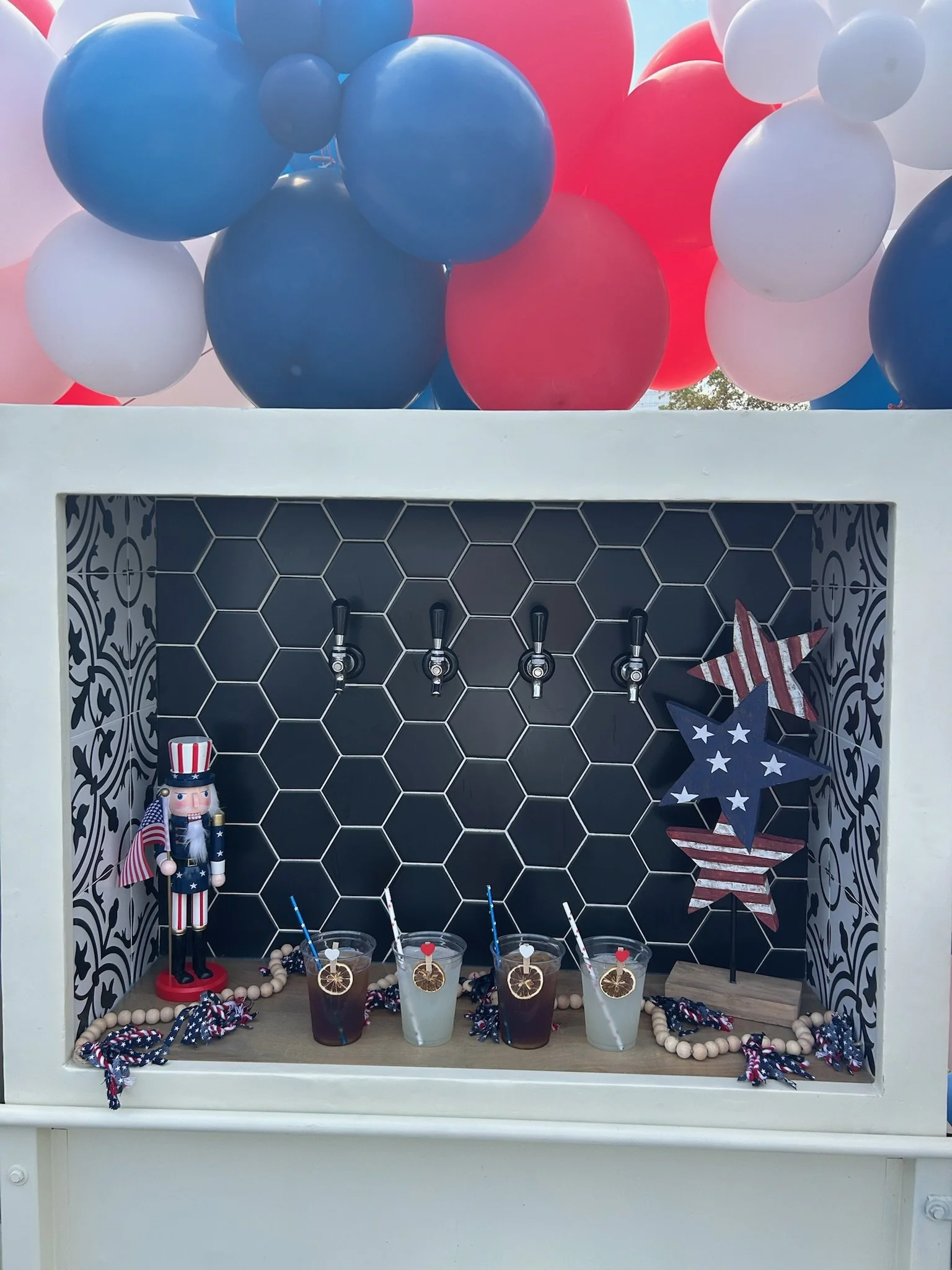 Fourth of July themed beverage station with four drinks, patriotic decorations, and balloons in red, white, and blue colors.