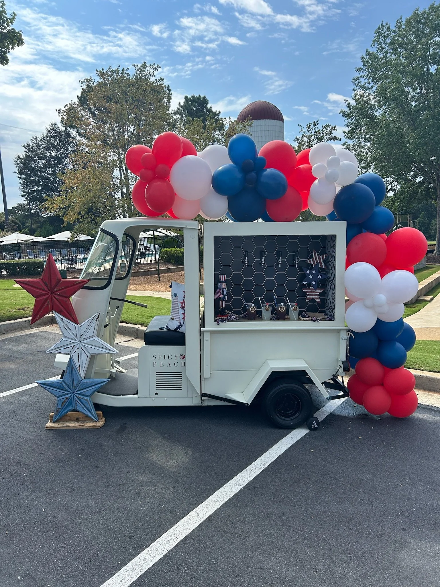 A small white food cart decorated with red, white, and blue balloons and stars for an American patriotic celebration, parked in an outdoor parking lot.
