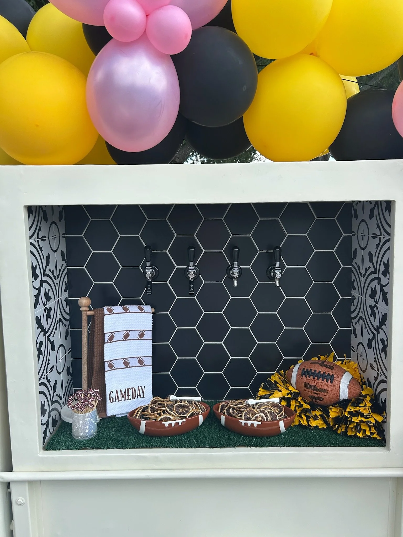 A game-day sports-themed display with black, pink, and yellow balloons above a white box containing small bowls of snacks, a football, and a small towel.