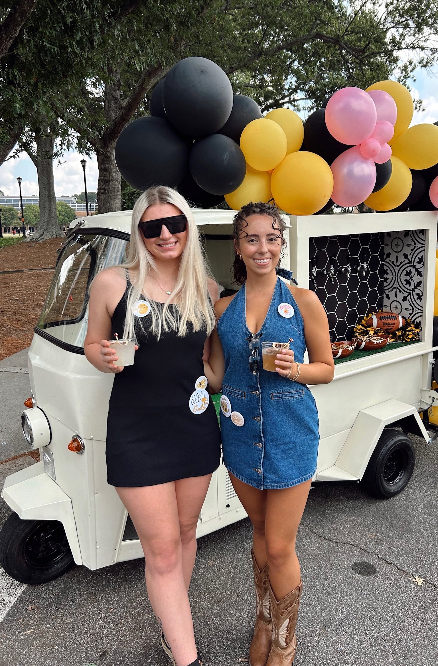 Two women standing in front of a small white vehicle decorated with black, yellow, and pink balloons, holding drinks and smiling at the camera.
