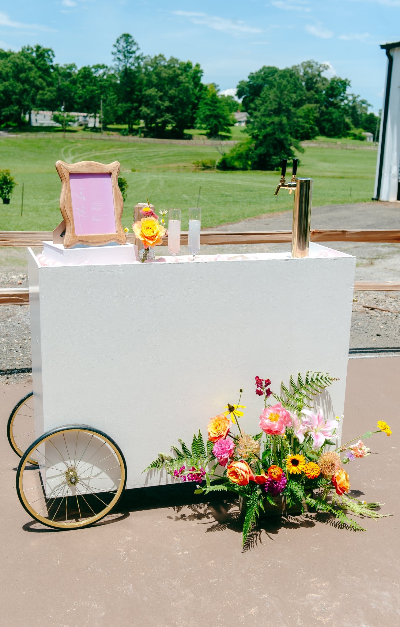 A white food cart decorated with flowers and a chalkboard sign, set outdoors on a sunny day with green fields and trees in the background.
