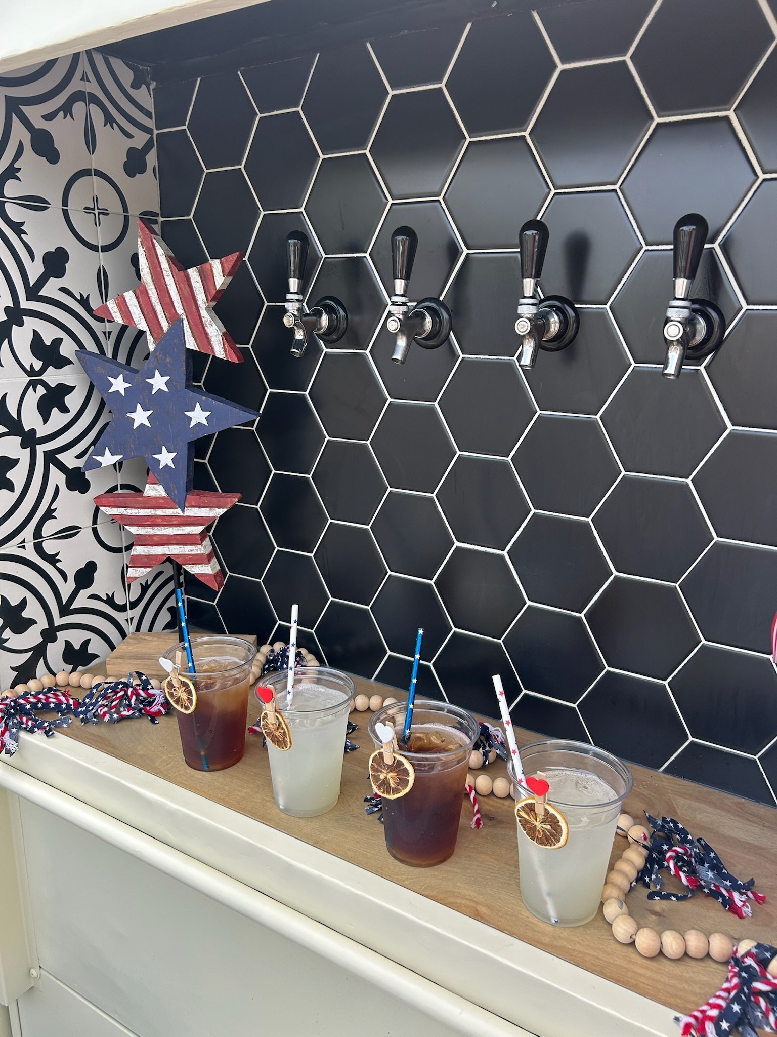 Patriotic-themed drinks with ice, garnished with dried citrus slices and decorative stir sticks, on a counter decorated with red, white, and blue star and stripe ornaments for Independence Day celebration.