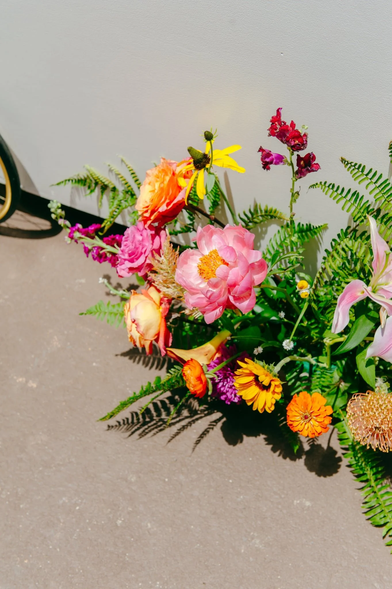 A colorful bouquet of flowers with pink, yellow, orange, purple, and red blossoms arranged with green fern leaves, casting shadows on a gray surface. A detached wheel with a yellow rim is partially visible in the upper left corner.