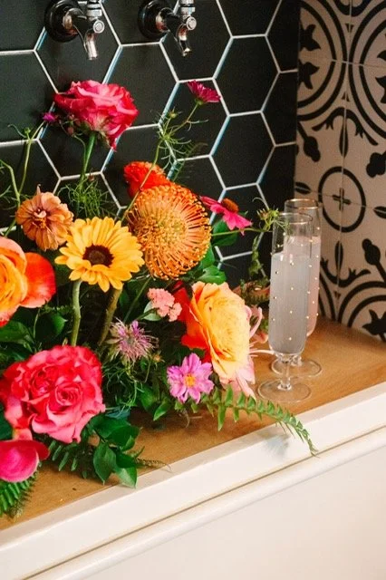 A colorful bouquet of flowers including pink roses, sunflowers, and other blooms in front of a black hexagon-tile wall with a faucet, along with a glass of sparkling water on a wooden surface.