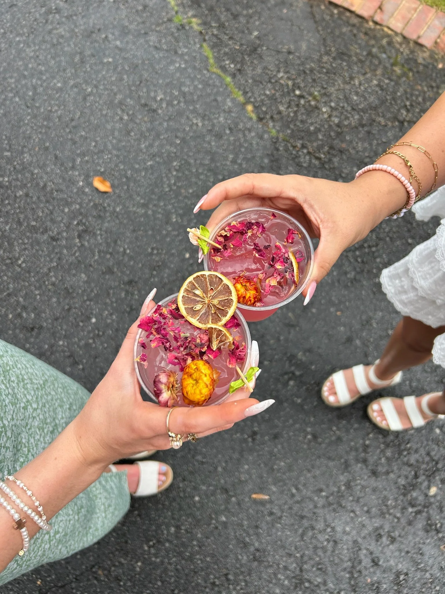 Two people holding drinks garnished with dried lemon slices, flowers, and petals, standing on a paved surface.
