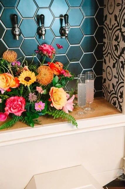 A floral arrangement with various colorful flowers, two glasses of pink drink, and three drink taps mounted on a teal-tiled wall.