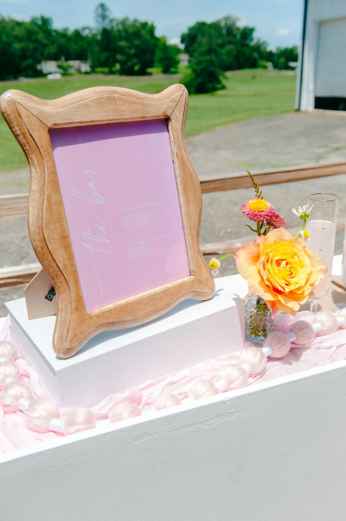 A pink framed sign with white text that appears to say 'I do' and some floral arrangements with pink and yellow flowers on a white table at an outdoor wedding or event setting.