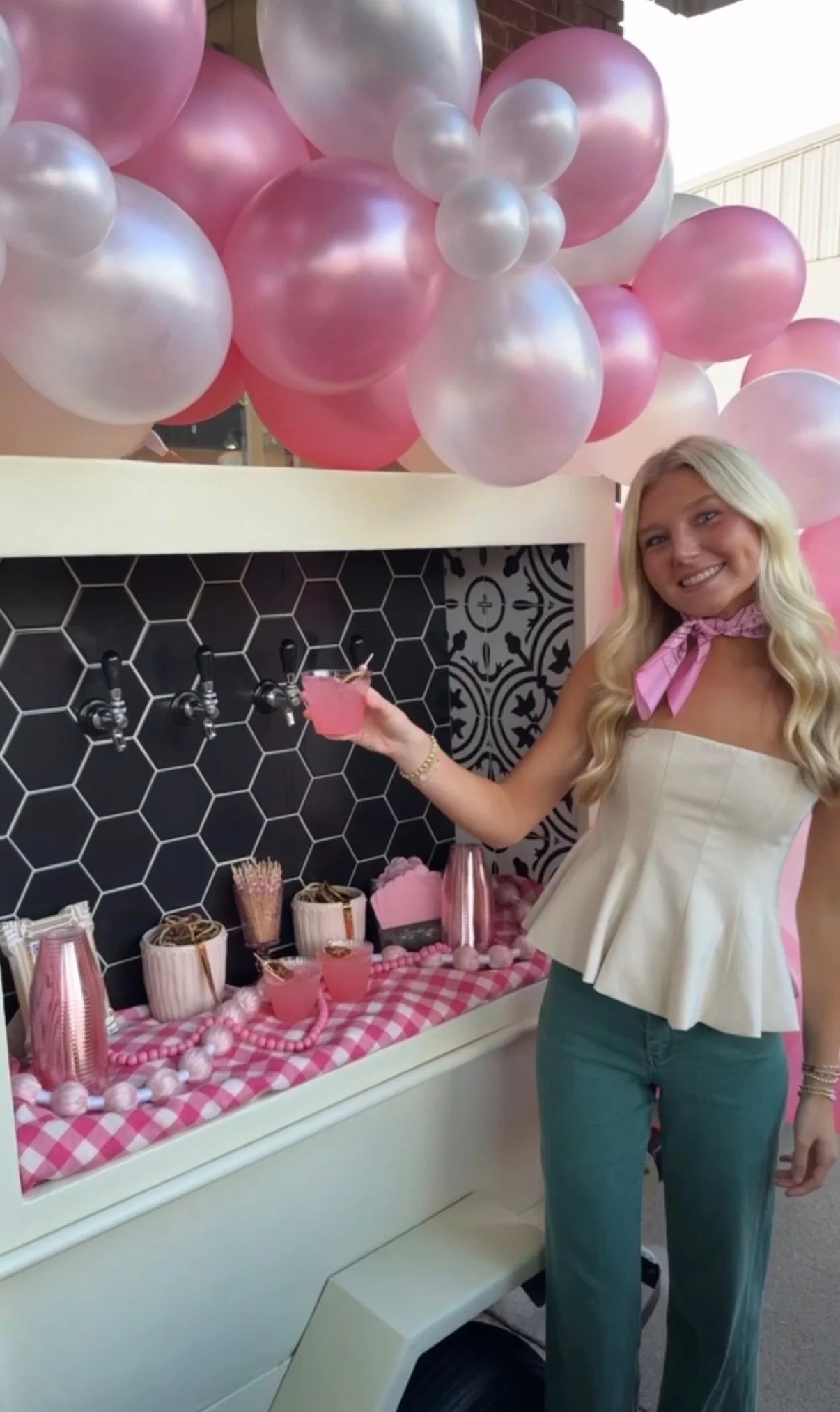 A woman with long blonde hair wearing a strapless beige top, blue jeans, a pink scarf, and jewelry, holding a pink drink in front of a beverage station decorated with pink and pearl-colored balloons.