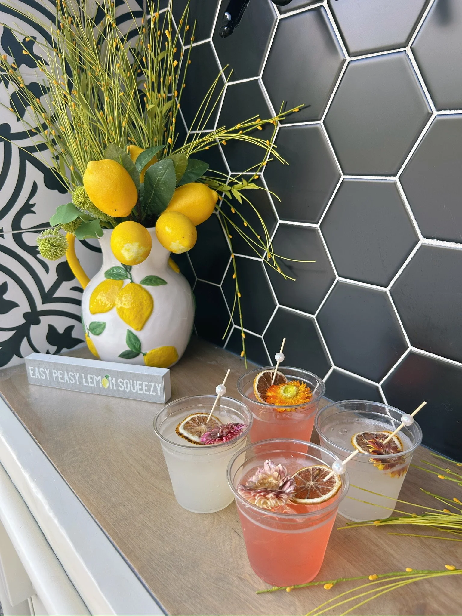 A decorative yellow lemon-themed vase with artificial lemons and green leaves, placed on a light wood surface. Next to it are three colorful drinks garnished with slices of dried lemon and edible flowers, each with a small skewer. The background feat