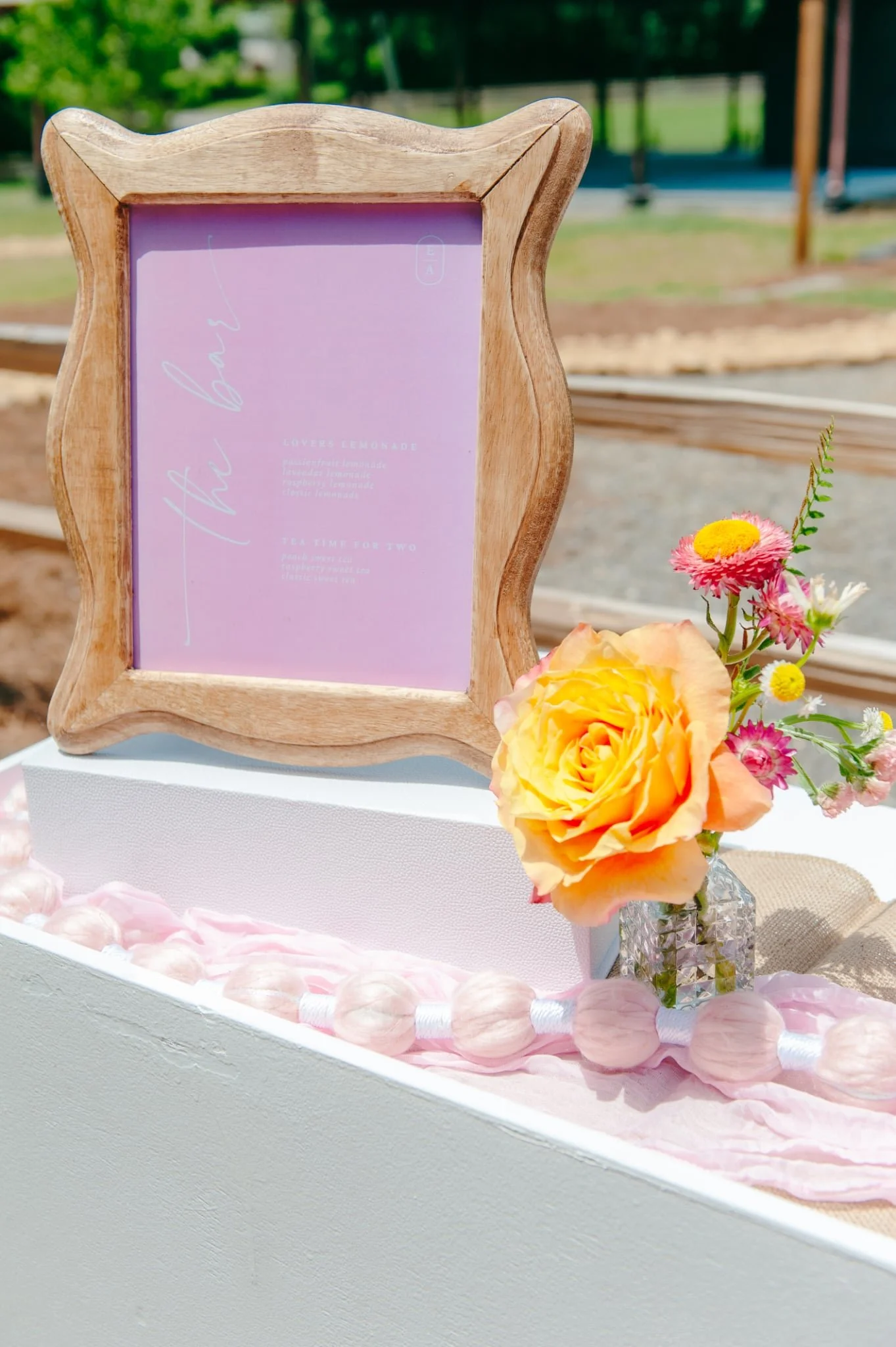Close-up of a wooden-framed lavender sign with white cursive text, beside a small glass vase with pink, orange, and white flowers, decorated with pink and white flower petals on a white surface outside.
