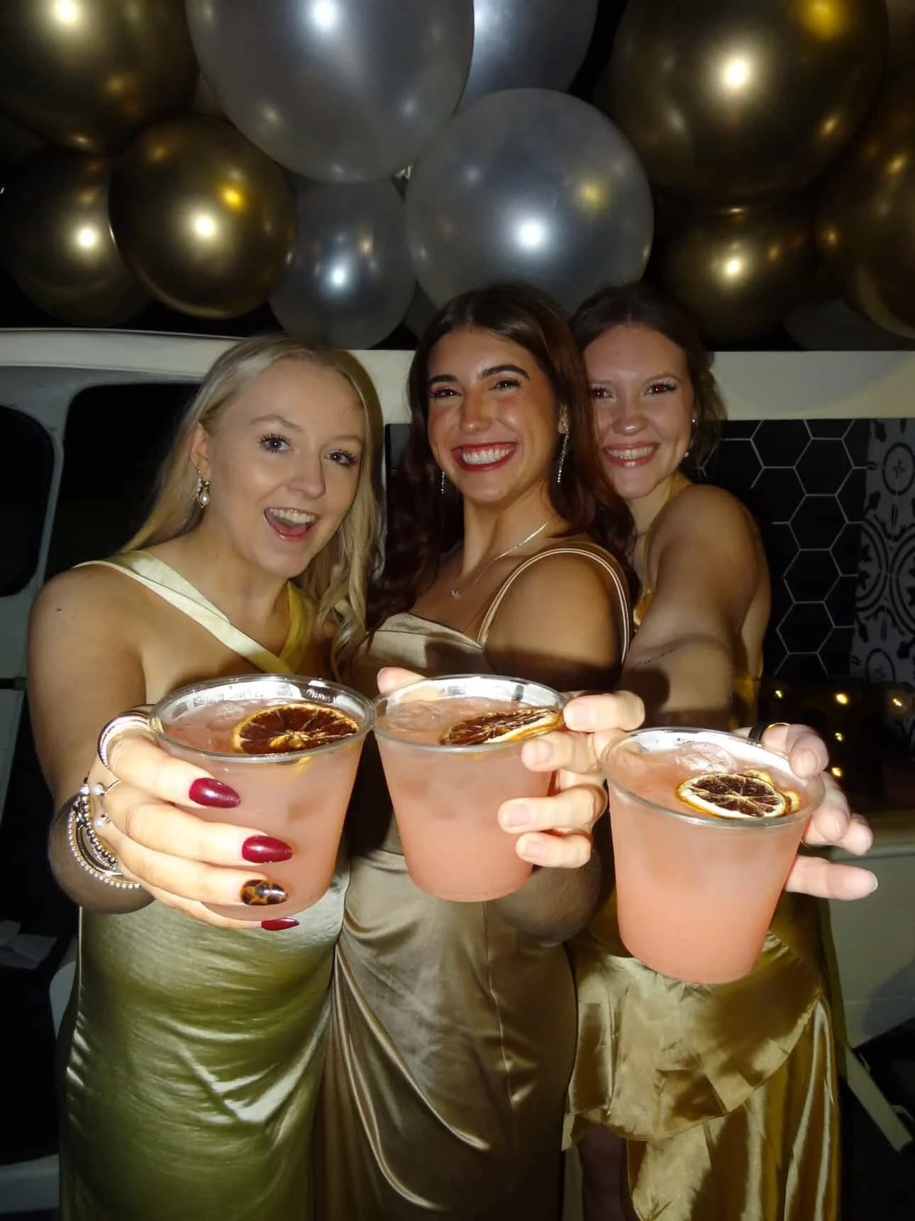 Three women in gold dresses celebrating with pink cocktails garnished with dried citrus slices, holding their drinks toward the camera at a party with black, gold, and silver balloons.