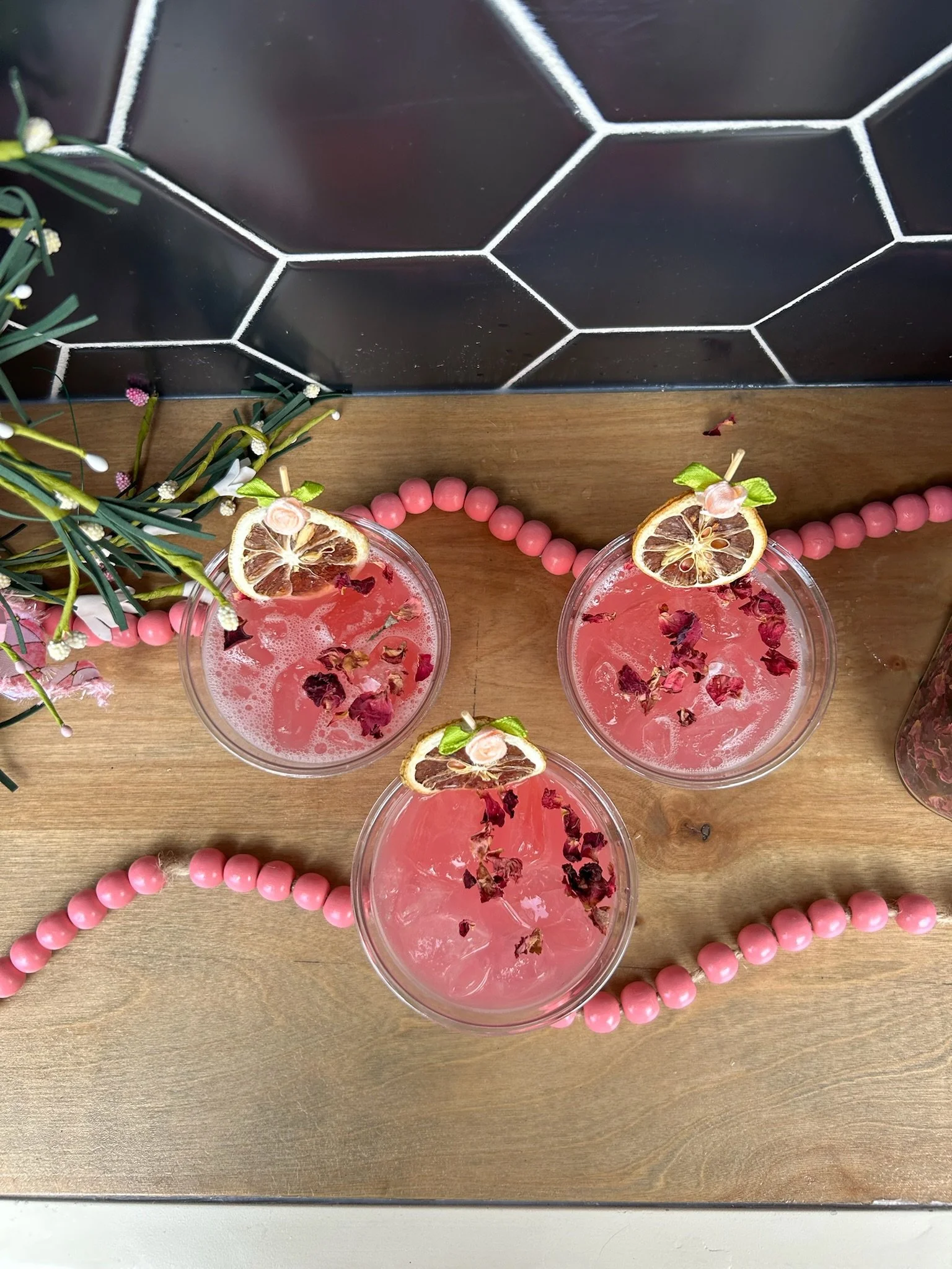 Three pink beverages garnished with dried lemon slices and rose petals, set on a wooden surface decorated with pink beads and a small floral arrangement, against a black tiled wall.