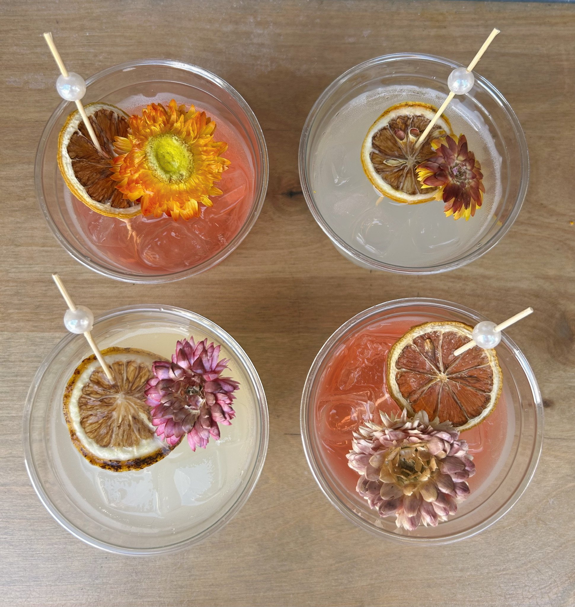 Four cocktails with dried lemon slices and edible flowers, served in clear glasses with skewers topped with pearls, on a wooden surface.