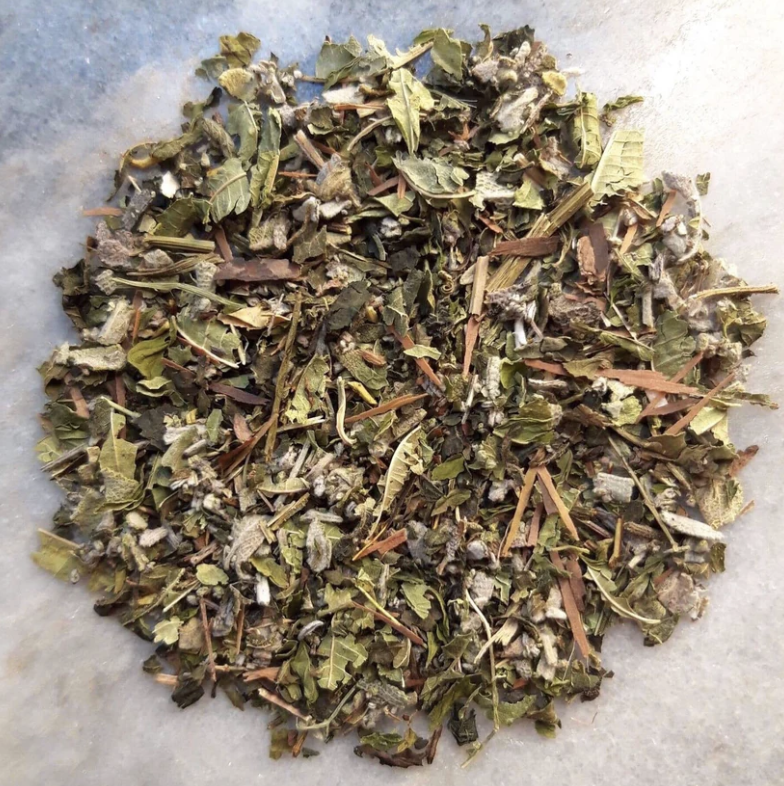 Dried herbal mixture with green leaves, small stems, and tiny flowers on a light surface.