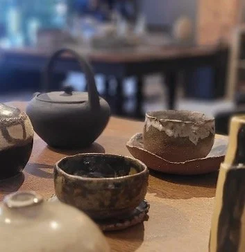 Close-up of several small, rustic pottery bowls and teapots on a wooden table in a cozy, cafe-like setting.