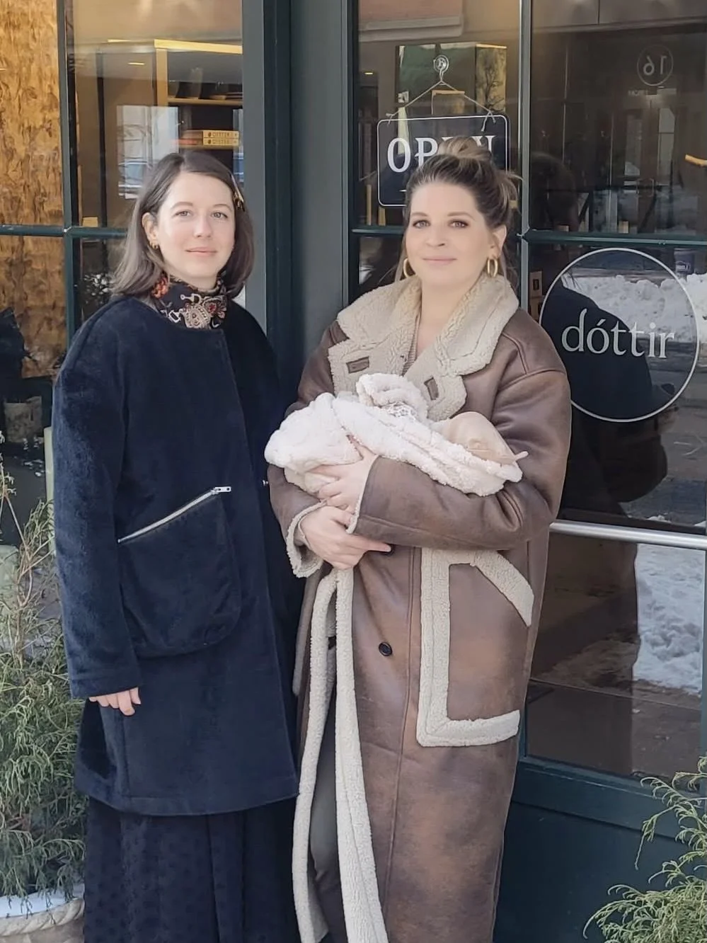 Two women stand outside a shop, one holding a newborn baby wrapped in a blanket, in front of a glass door with signs that say 'OPEN' and 'dóttir'.