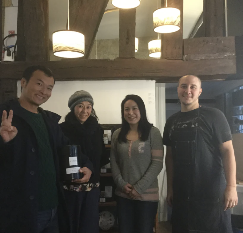 Group of four people standing together indoors, with some holding a container, smiling at the camera, in a cozy setting with warm lighting.