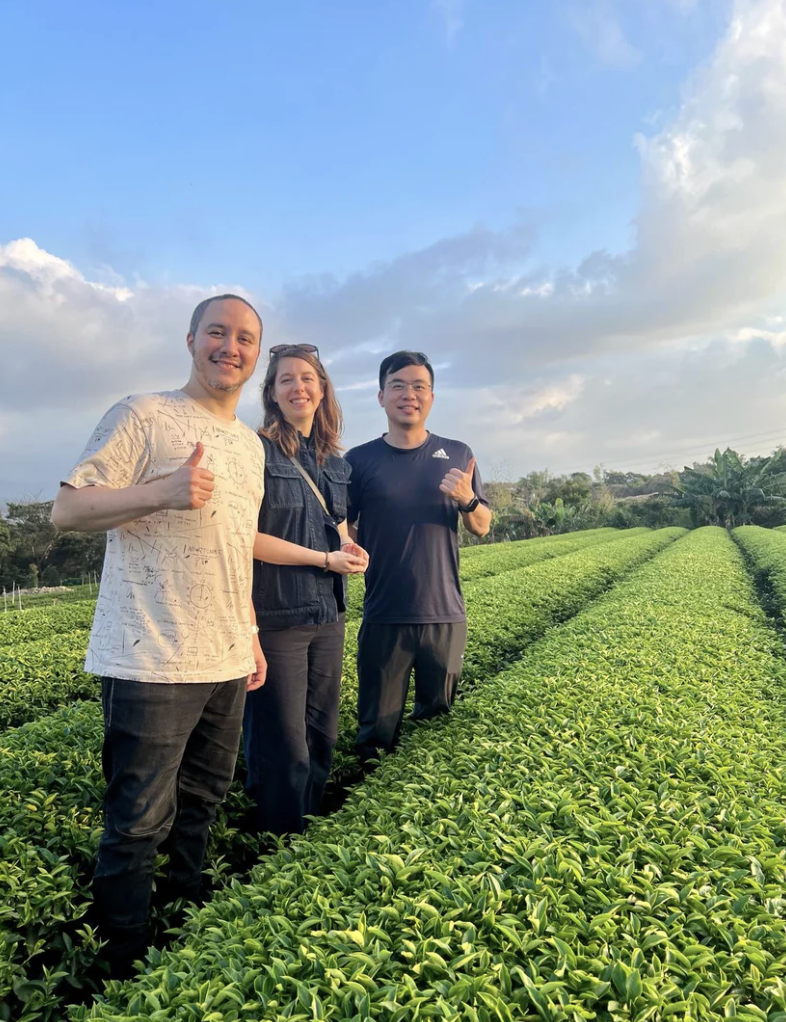 Three people standing in a lush green field with rows of plants, under a partly cloudy sky.