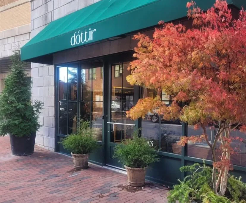storefront with teal awning labeled 'dóttir', large glass windows, brick sidewalk, greenery in pots, and a red-orange autumn tree in front.