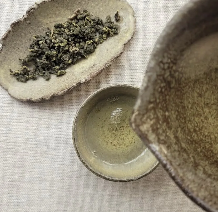 A ceramic bowl of hot water, an oval dish with dried tea leaves, and a ceramic teapot used for brewing loose leaf tea, on a light fabric surface.