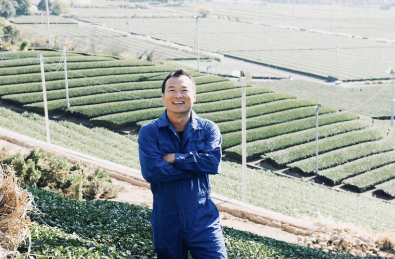 Man smiling with arms crossed standing in a tea plantation with rows of tea plants and hilly landscape in the background.