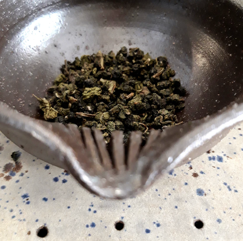 Close-up of a metal spoon with dried green and black herbs on a speckled countertop.