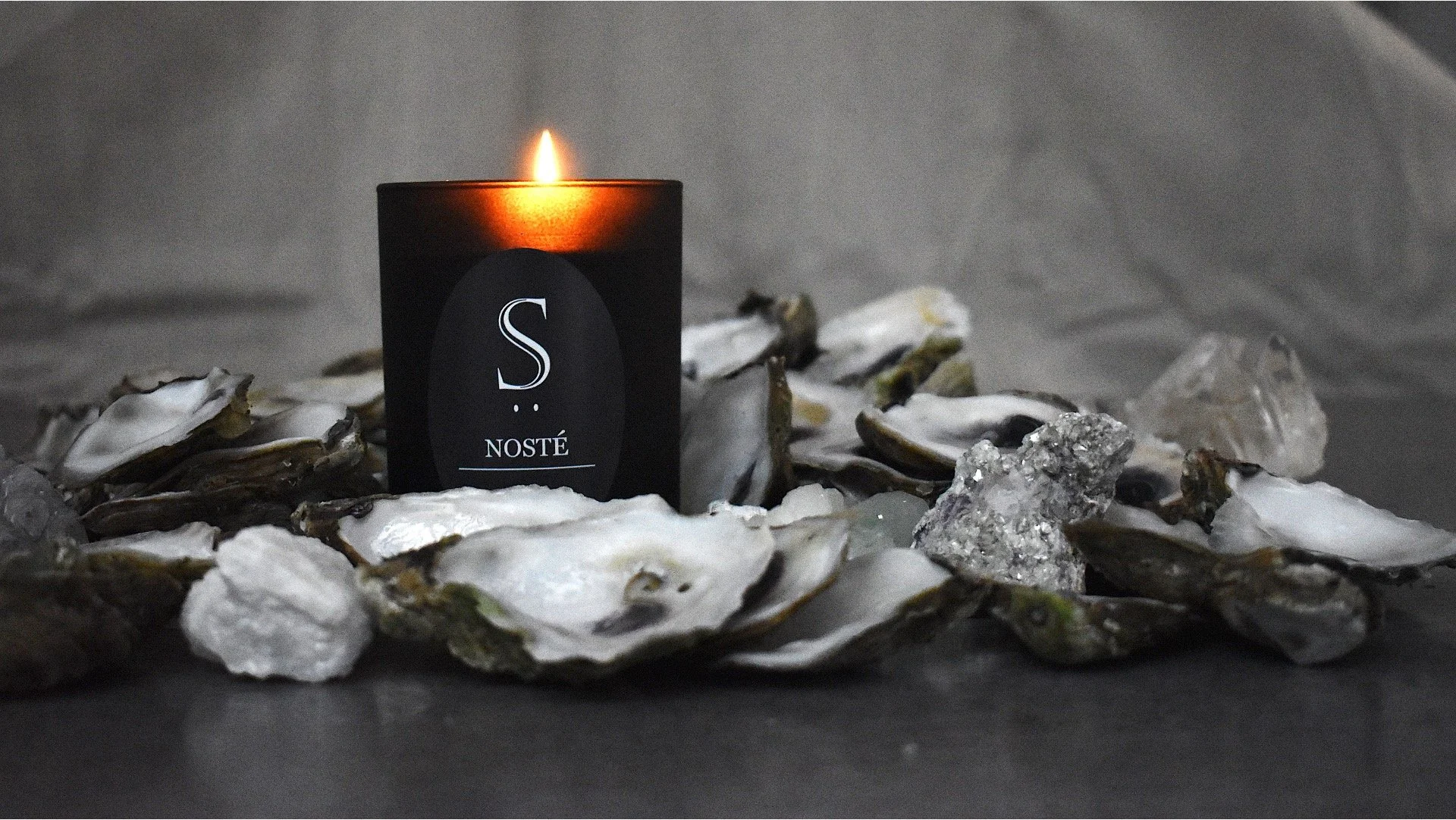 A lit black candle with a label reading 'S NOSTÉ' surrounded by oyster shells and ice on a dark surface.