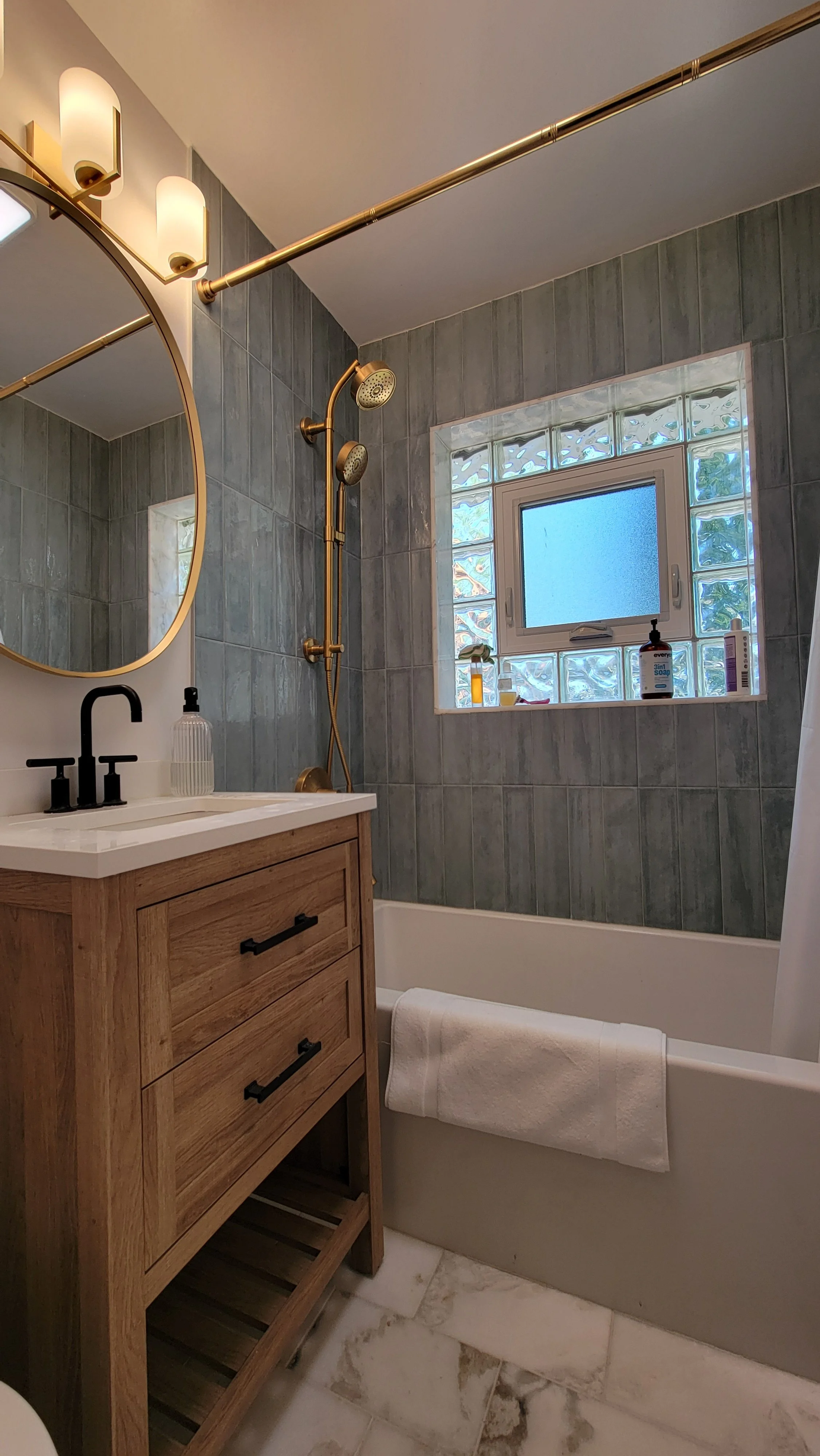 A bathroom with green tiled walls, a bathtub with a towel, a wooden vanity with a sink, a round mirror, a window with glass blocks, and a gold shower fixture.