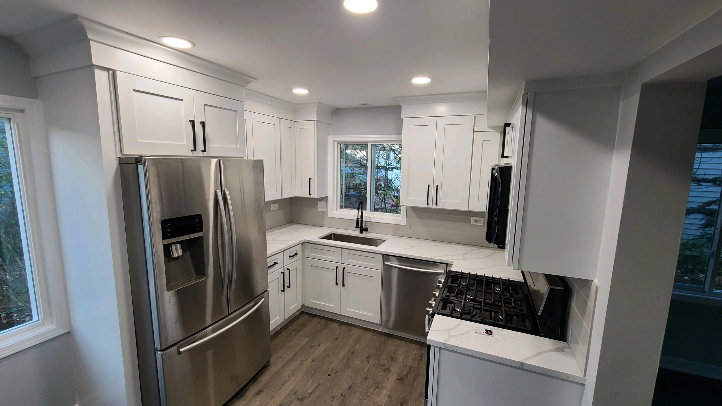 Modern kitchen with white cabinets, stainless steel refrigerator, marble countertops, black handles, and a window above the sink.