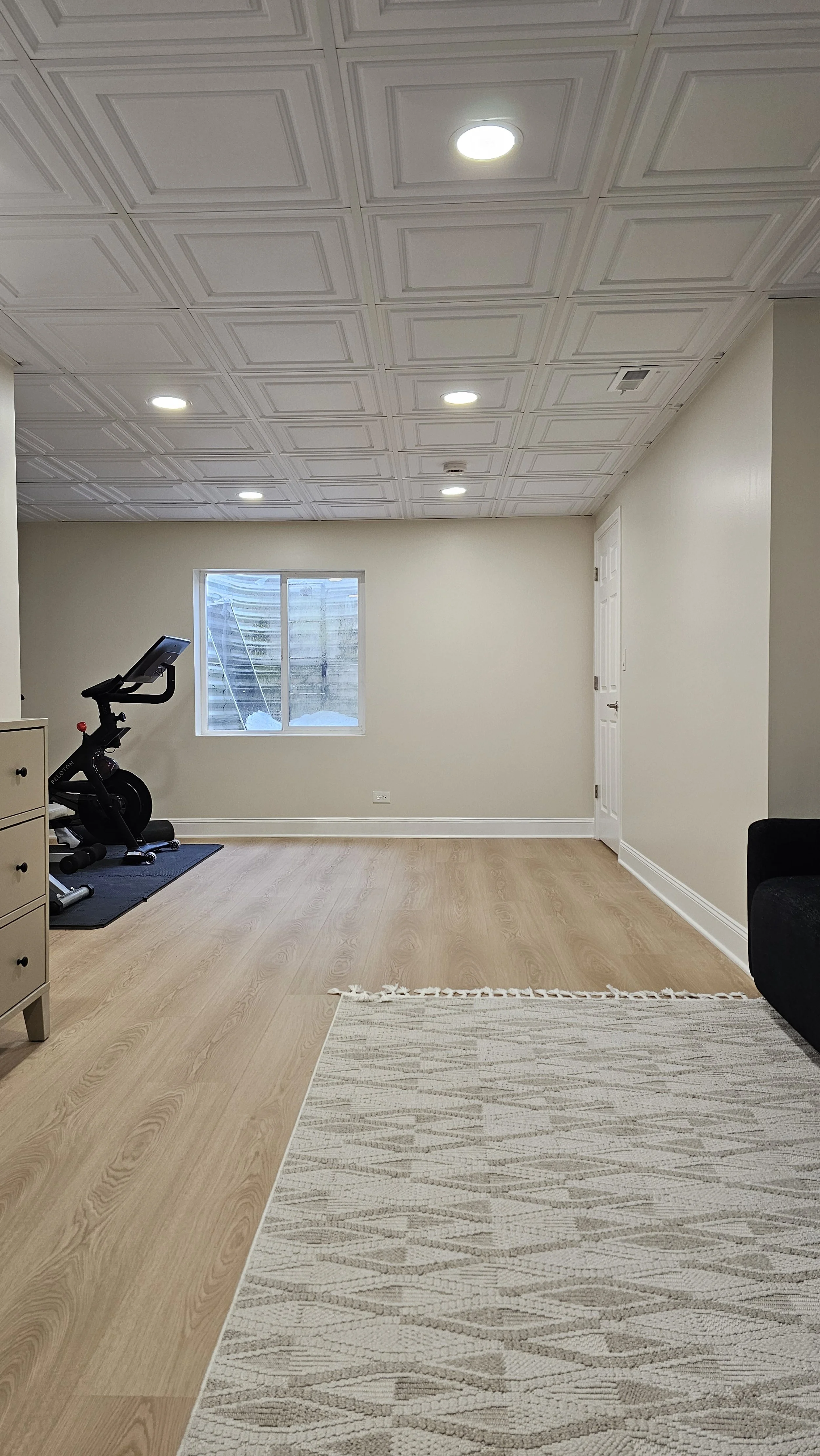 Empty room with wooden floor, a window with snow outside, exercise bike, dresser, and a black sofa.